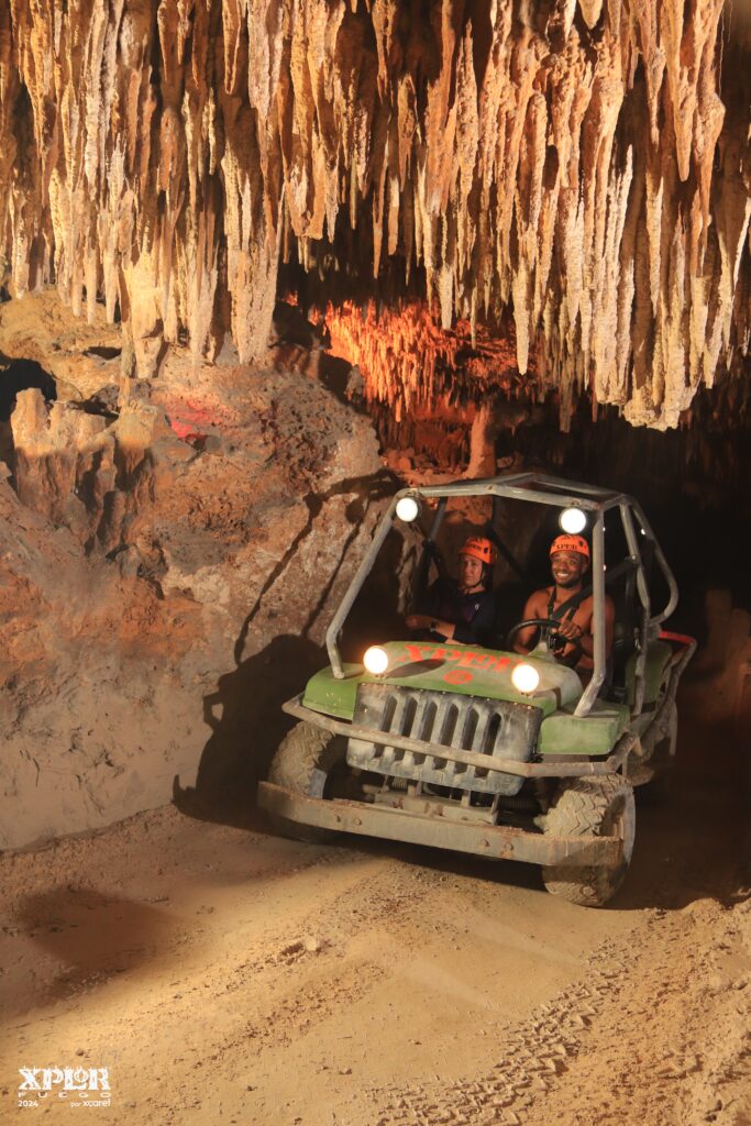 wo people wearing helmets drive a green ATV through a cave filled with stalactites, illuminated by warm orange lights, with rough cave walls surrounding them.