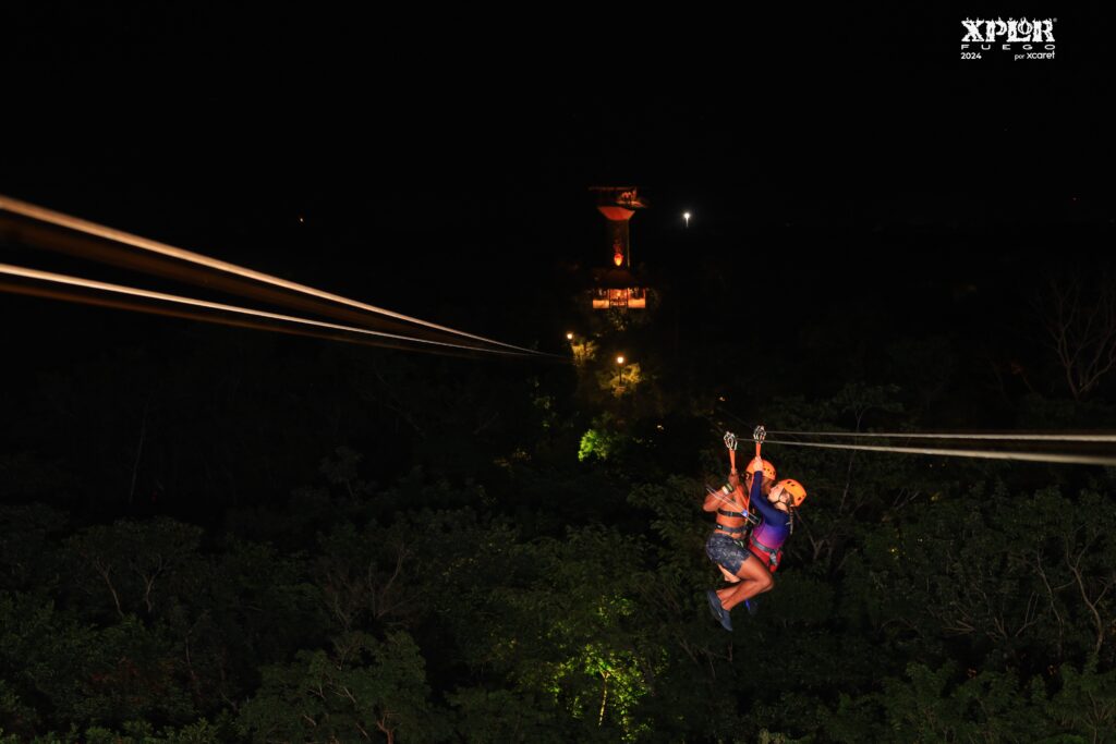 Two people zip-lining at night through the jungle at Xplor Fuego, an adventure park in Mexico. They are wearing helmets and harnesses, gliding along illuminated cables above dense green foliage. In the background, a fire-lit observation tower adds to the dramatic nighttime atmosphere.