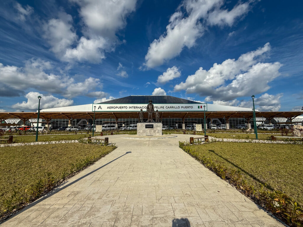 A wide-angle view of Tulum International Airport (Aeropuerto Internacional Felipe Carrillo Puerto) under a bright blue sky with scattered clouds. The modern terminal building features a sleek, eco-chic design with a wooden canopy structure. A stone pathway leads to a statue in front of the entrance, surrounded by neatly maintained green landscaping.