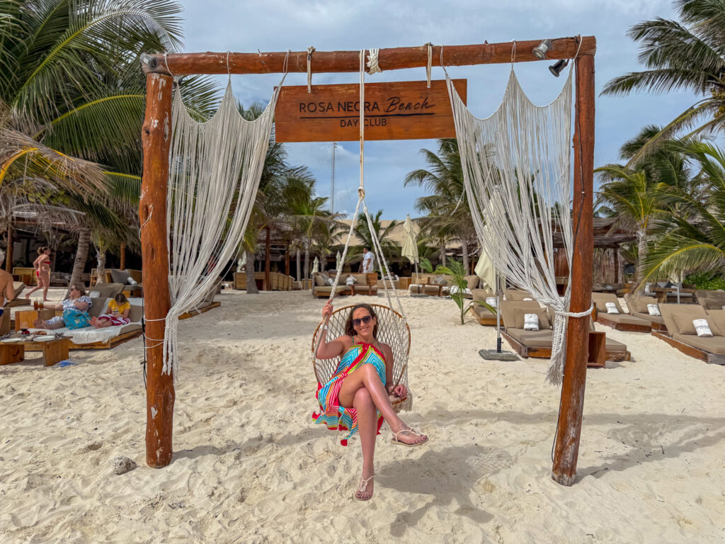 A woman in a colorful dress sits on a hanging chair swing framed by a wooden arch decorated with white macrame drapes. The sign above reads "ROSA NEGRA Beach Day Club," with sandy beach loungers, palm trees, and other guests relaxing in the background.