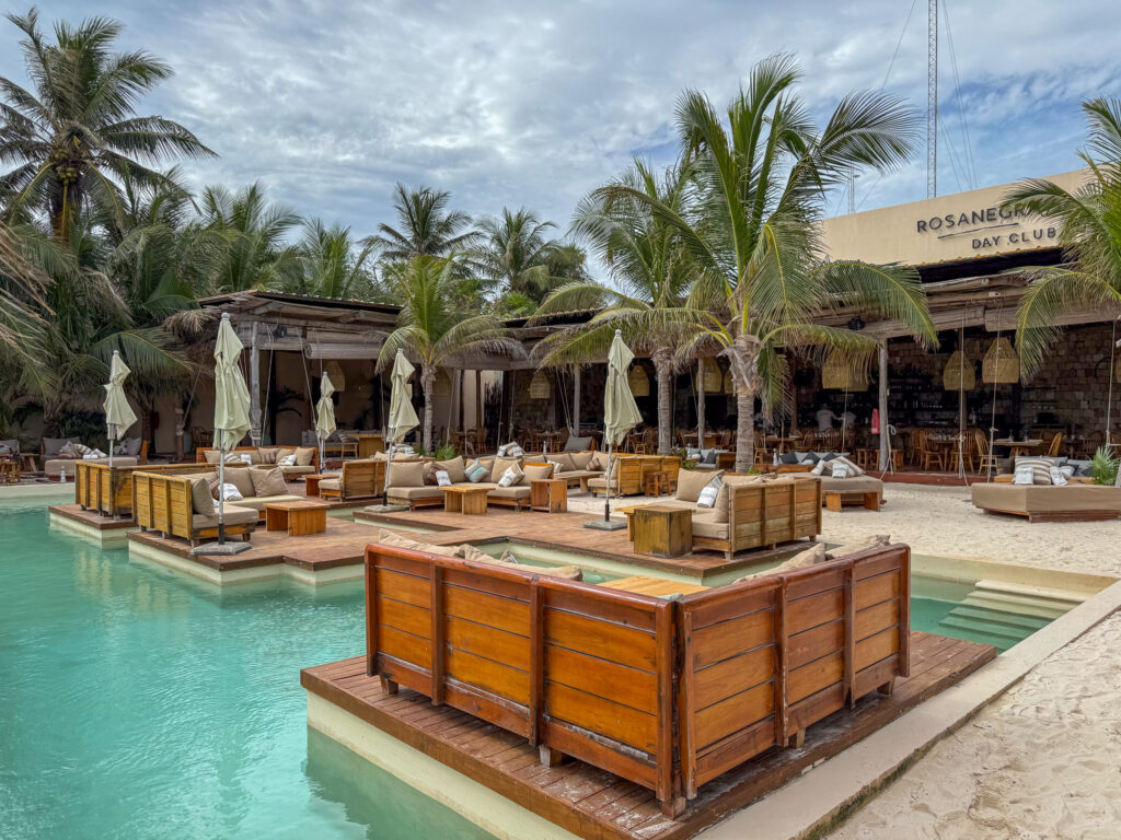 A tropical beach club pool surrounded by wooden platforms with plush beige seating, umbrellas, and palm trees. The background features a rustic open-air bar and dining area with wicker light fixtures and wooden chairs, with "ROSANEGRA Day Club" displayed on the building facade.