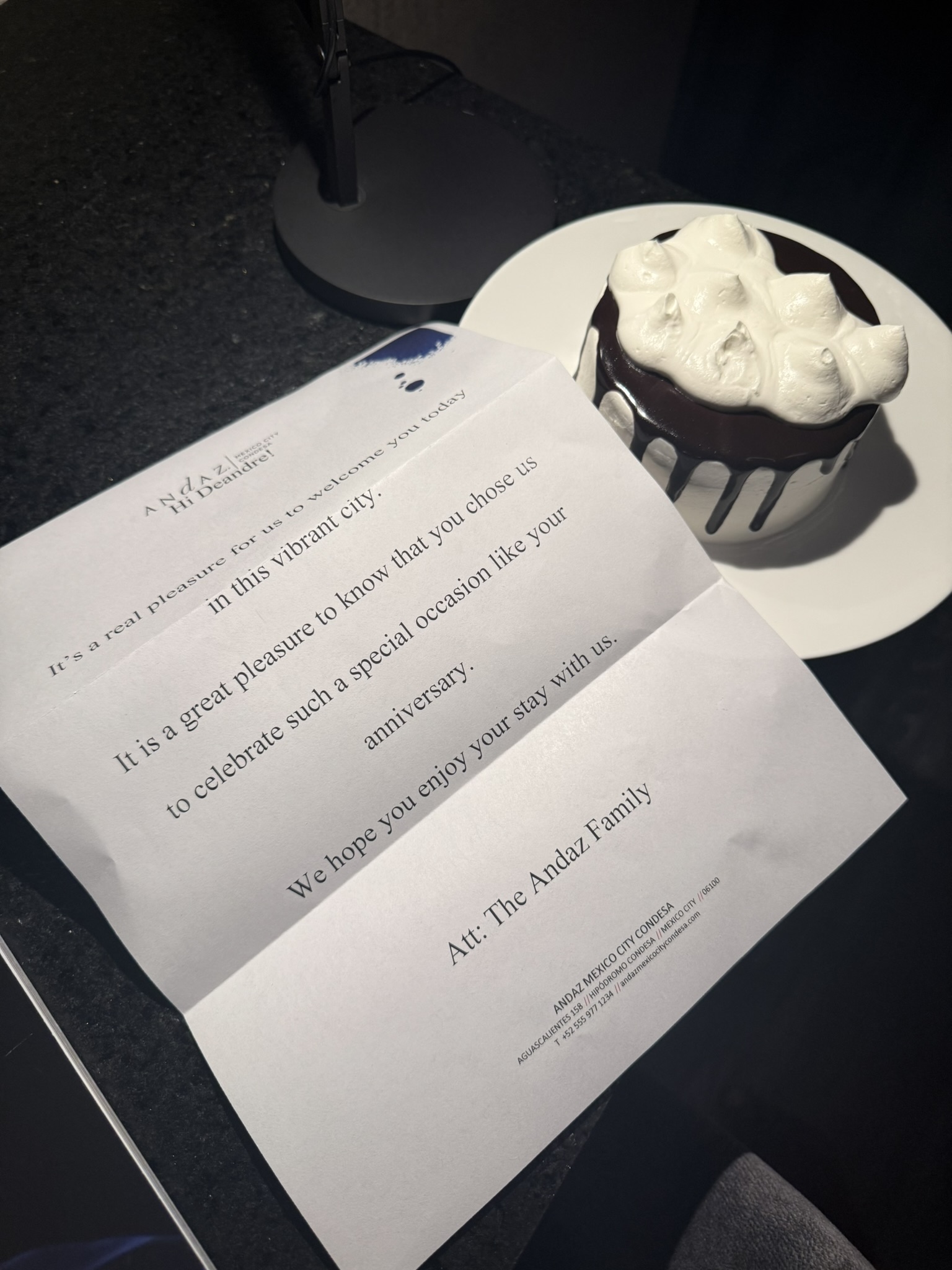 A welcome note from Andaz Mexico City Condesa addressed to "DeAndre," placed on a dark countertop next to a small celebratory cake. The cake has a white whipped topping with chocolate drizzle, sitting on a white plate, as part of a special gesture for an anniversary celebration.