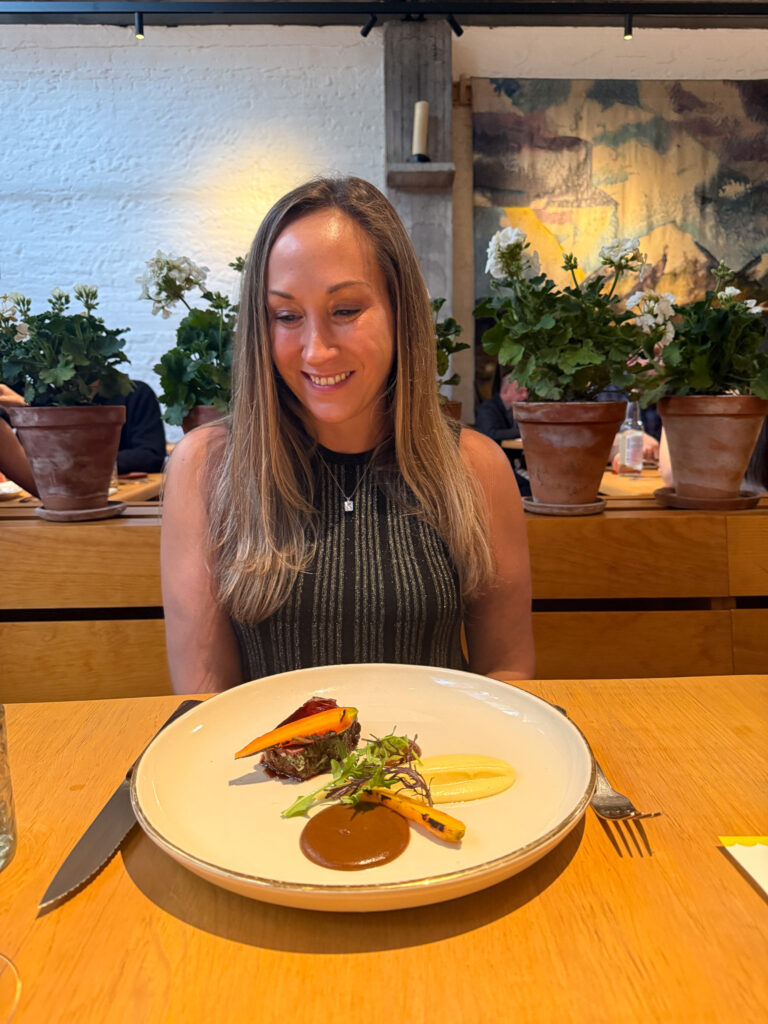 A woman sitting at a modern fine-dining restaurant, smiling at an elegantly plated dish featuring a gourmet cut of meat, sauce, and vegetables. The restaurant has a warm, minimalist ambiance with potted plants and artistic decor.