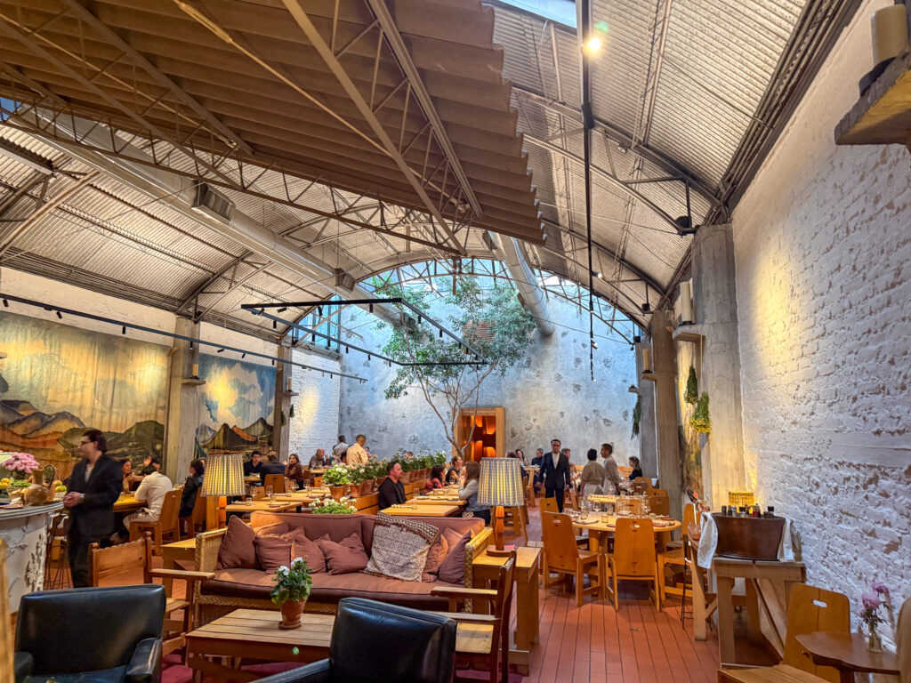  A beautifully designed restaurant with a spacious, open-air industrial-style interior featuring a high, arched metal ceiling, exposed white brick walls, and large murals of scenic landscapes. The space is filled with warm lighting, wooden furniture, and cozy seating areas with plush cushions. Guests are dining at tables, while staff move around providing service, creating a lively yet intimate ambiance