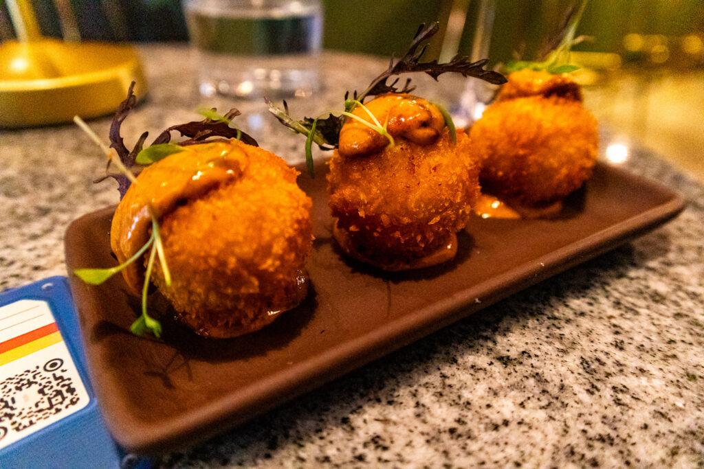 close-up of three crispy golden croquettes, garnished with microgreens and drizzled with a rich sauce, served on a dark ceramic plate in a dimly lit upscale restaurant.