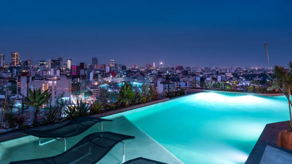 An illuminated infinity pool on the rooftop of Andaz Mexico City, overlooking the skyline at dusk, with lounge chairs and lush greenery surrounding the space.