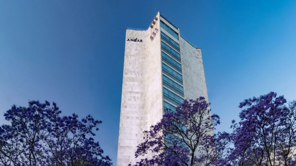 A tall, modern Andaz Mexico City hotel building with a white facade, set against a clear blue sky, framed by lush jacaranda trees with vibrant purple blooms.