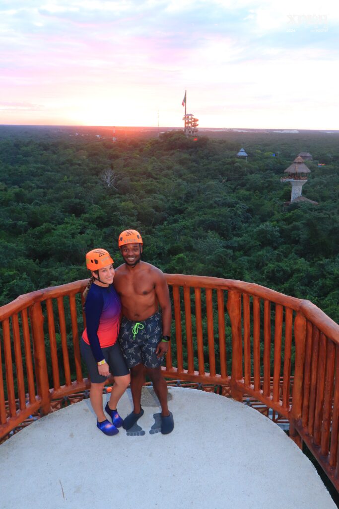 A couple in helmets stands on a circular wooden platform, smiling with their arms around each other. The background features lush green jungle, observation towers, and a sunset sky blending pink, orange, and blue hues.