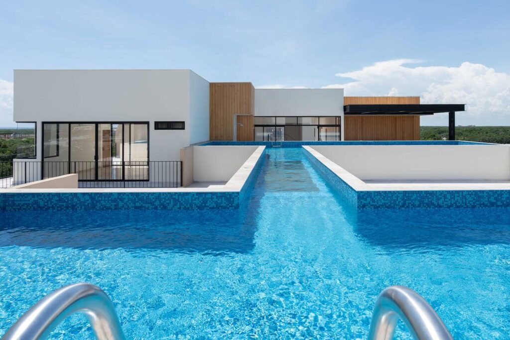 A rooftop swimming pool with clear blue water, surrounded by white and wooden modern architecture, with a view of lush greenery in the distance under a bright blue sky.