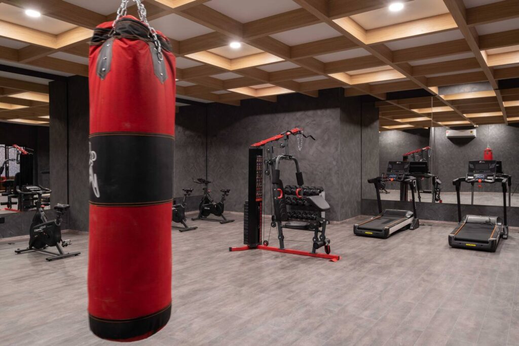 A modern gym with a red punching bag hanging in the foreground, stationary bikes, treadmills, and strength training equipment in the background. The space has dark gray walls, wood-paneled ceilings, and mirrored walls.