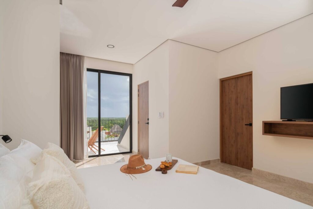 A bright bedroom with a white bed facing a mounted TV, sliding glass doors leading to a private balcony with lounge chairs, and two wooden doors, all with natural light streaming in.