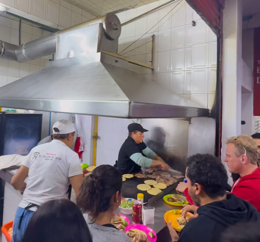 A bustling taqueria in Mexico City with a cook grilling meat and heating tortillas on a flat-top grill. Customers are gathered around the counter, enjoying freshly made tacos. A large metal ventilation hood hangs above the cooking area.