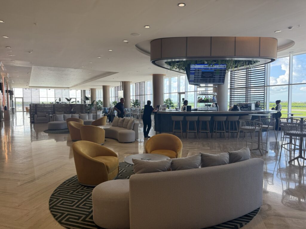 Modern open-air lounge interior at the Punta Cana VIP Lounge featuring a circular bar, flight departure screen, and stylish seating with large windows overlooking the runway.