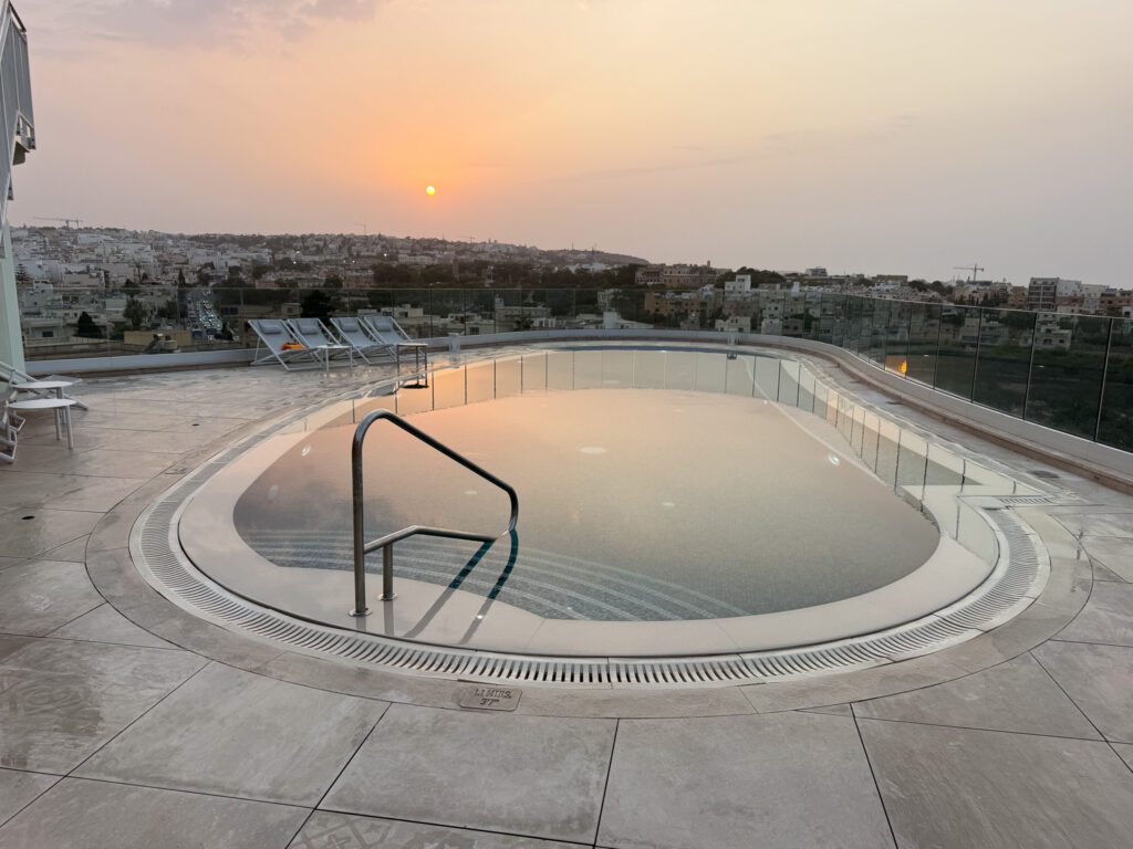 Sunset reflected in the rooftop pool, with curved entry steps and city buildings beyond the glass railing