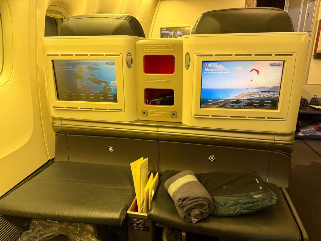 Two seatback entertainment screens above broad ottomans in Turkish Airlines 777 business class.